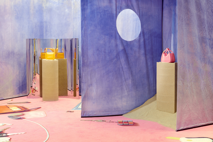 Installation mit violetten Wänden, rosa Teppich und gelben Säulenhocker auf welchen pinke und gelbe Handtaschen präsentiert werden.