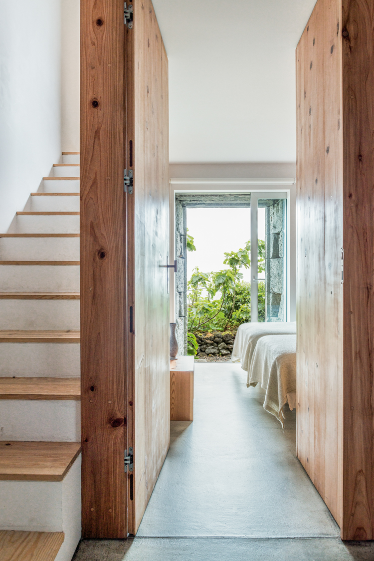 Wohnraum mit Gang mit Blick auf offene Fensterfront mit Blick in den Garten, links führt eine Treppe mit Holzstifen in den oberen Stock.