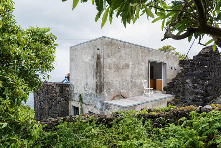 Blick durchs Gebüsch auf ein kubus-artiges Haus mit alten Steinmauern.