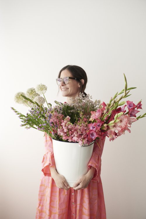 Ulrike Krucker von Blüemlisex steht mit rosa Kleid vor weisser Wand und hält einen weissen Topf mit Blumen vor ihrem Oberkörper.
