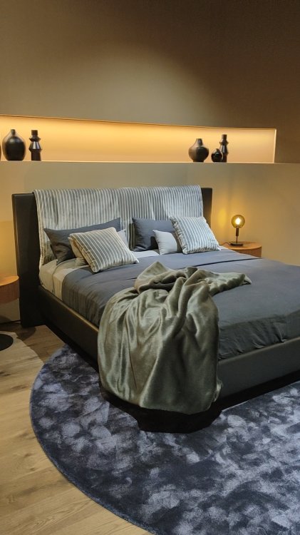 Ein Foto eines Bettes mit grauen Überzug, welches auf einem Teppich steht.