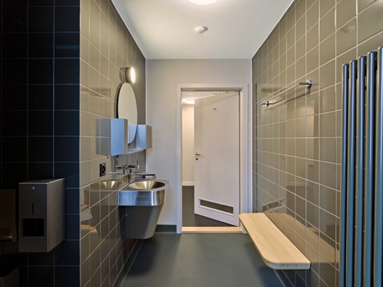 Sanitärraum mit Edelstahl-Doppelwaschbecken, Spiegeln, grauen Wandfliesen, Holzbänkchen und Heizkörper.