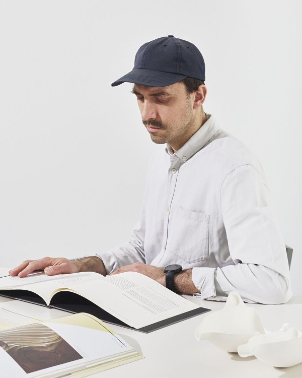 Designer Tomáš Král mit Kappe liest an einem Tisch, umgeben von Büchern und hellen Objekten.