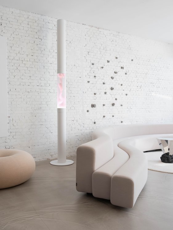 Hohe weisse Säulenlampe mit rosa fliessender Lava in einem hellen Raum mit weisser Backsteinwand und Designer-Sofa.