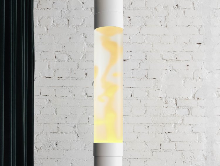 Nahaufnahme einer hohen weissen Säulenlampe mit sanft gelb-oranger Lava vor einer weissen Backsteinwand.