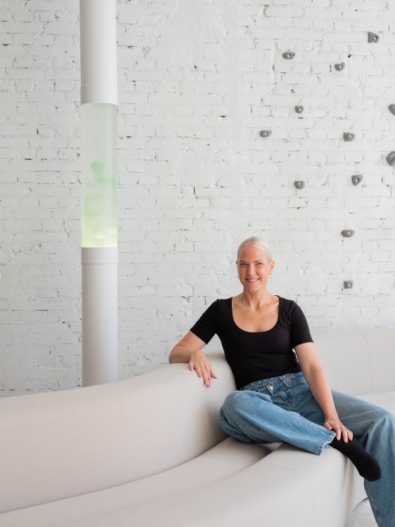 Sabine Marcelis mit kurzem blondem Haar sitzt auf einem beigen Sofa vor einer hohen Lavalampe mit grünlicher Lava und weisser Backsteinwand.