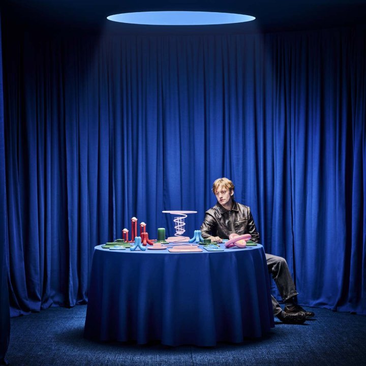 Designer Gustaf Westman sitzt an einem Tisch mit bunten Prototypen seiner IKEA-Kollektion vor blauem Hintergrund.