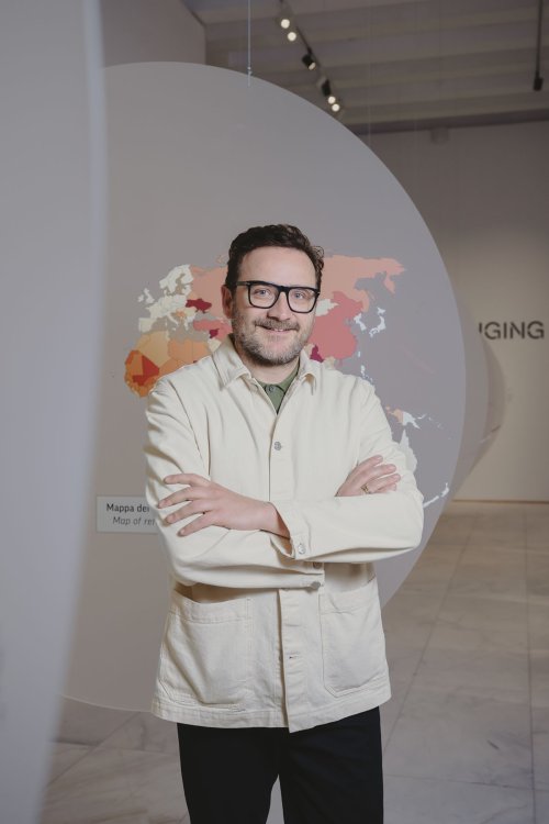 Porträt von Marco Sammicheli in einer Ausstellungssituation mit grafischer Installation im Hintergrund.