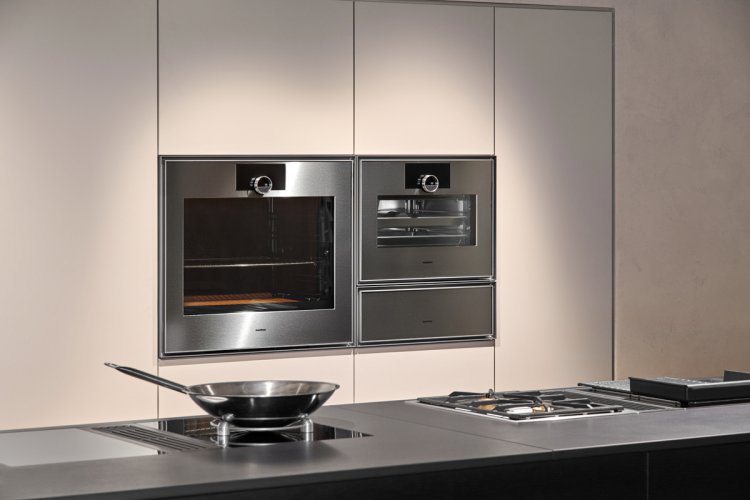 Backofen und Steamer in einer hellbeigen Küchenzeile integriert. im Vordergrund eine Herdplatte mit einer Pfanne.