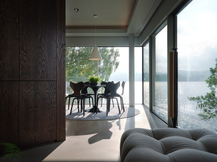 Heller, moderner Essbereich mit rundem Tisch und vier Stühlen vor bodentiefen Fenstern; Blick auf einen See und Bäume, Sonnenlicht fällt in den Raum und wirft lange Schatten.