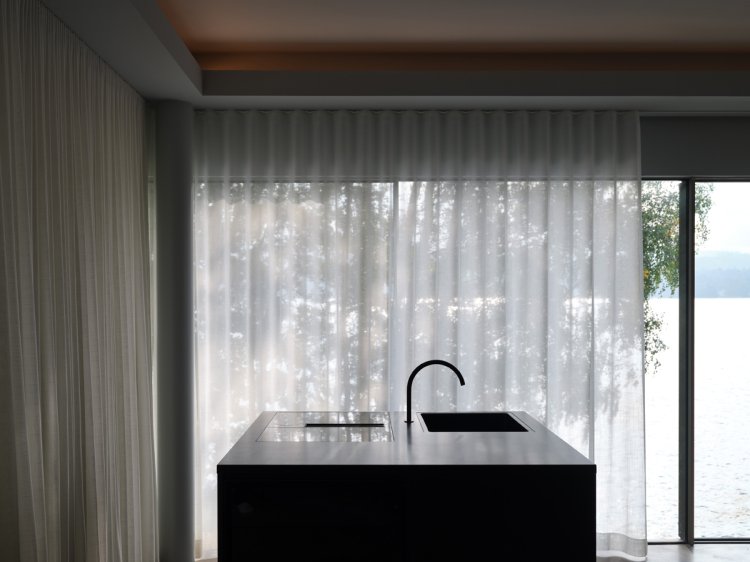 Blick auf eine Kochinsel, dahinter ein bodentiefes Fenster, halb von einer Gardine bedeckt. Im Hintergrund Wasser und Laubbäume.