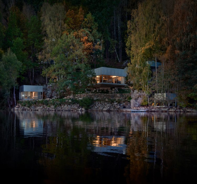 Mehrere moderne, kubische Häuser mit großen Glasfronten liegen am bewaldeten Ufer eines ruhigen Sees; warme Innenbeleuchtung spiegelt sich im dunklen Wasser, umgeben von herbstlich gefärbten Bäumen und Felsen.