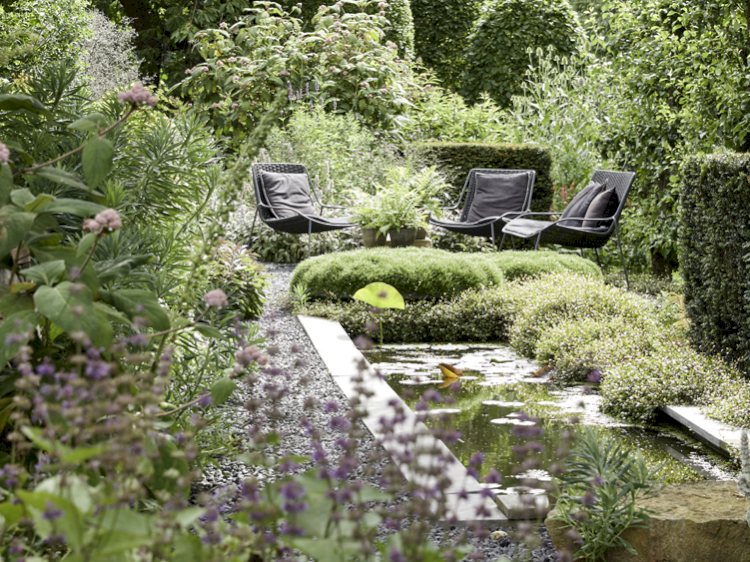 Drei filigrane graue Gartensessel inmitten eines Gartens, umgeben von Hecken, Gräsern, Pflanzen und Bäumen. Davor ein länglicher Seerosenteich.