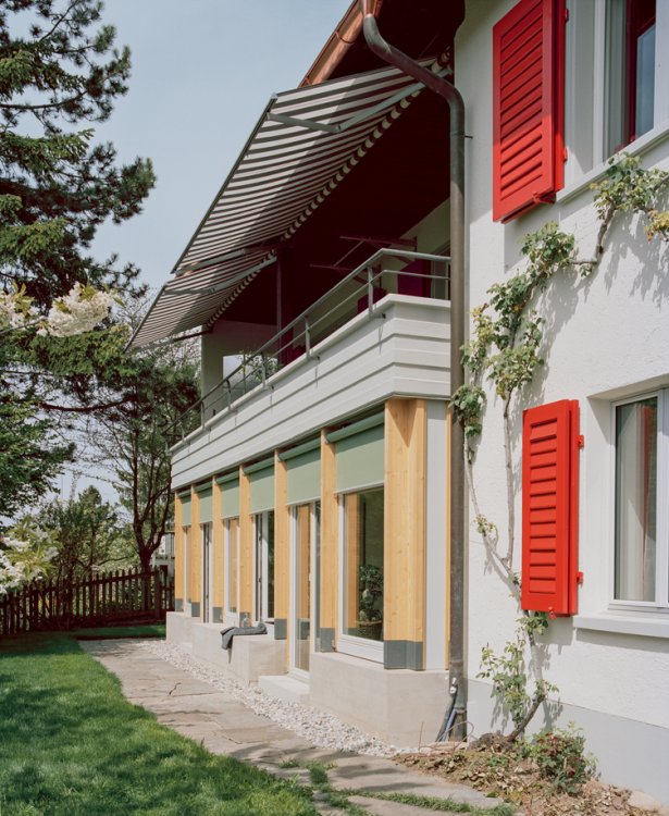 Modernisiertes Wohnhaus von 1953 mit verglastem Anbau unter den Bestands-Balkon mit Markise, aufgenommen vom Garten aus.