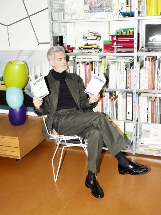 Ein Foto des Designers Alfredo Häberli, in welchem er auf einem Stuhl sitzt und in zwei Bücher hineinschaut.