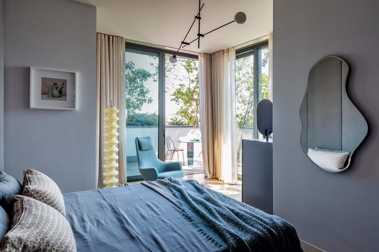 Ein Foto eines Schlafzimmer. Zu sehen ist ein grosses Bett mit blauer Decke. Vis a Vis vom Bett hängt ein abstrakter Spiegle an der Wand.