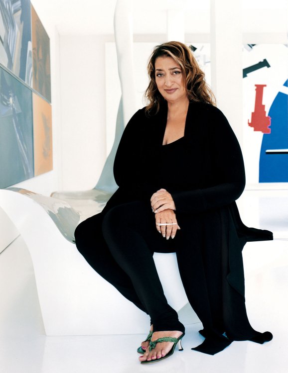Die Architektin und Designerin Zaha Hadid sitzt in einem schwarzem Outfit auf einem ihrer Stuhlentwürfe inmitten einer weissen Galerie.