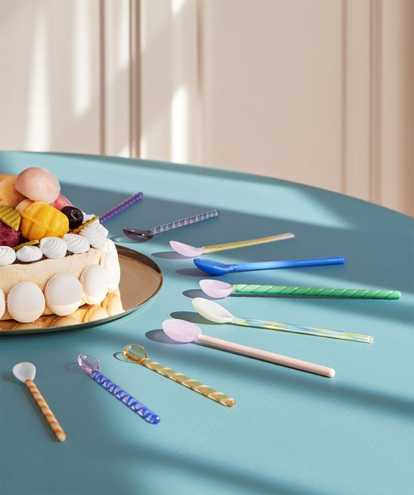 Ein üppiger Obstkuchen, um den unterschiedlich farbige und gemusterte Glaslöffel von HAY angeordnet sind, steht auf einem hellblauen, runden Tisch.