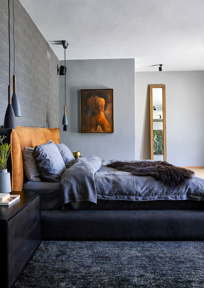 Ein grau gehaltenes Schlafzimmer mit einem Bett mit orangem Kopfteil und grauer Bettwäsche steht links an der Wand, im Hintergrund ist ein abstraktes Bild in Orangetönen sowie ein schmales, hohes Fenster zu sehen.