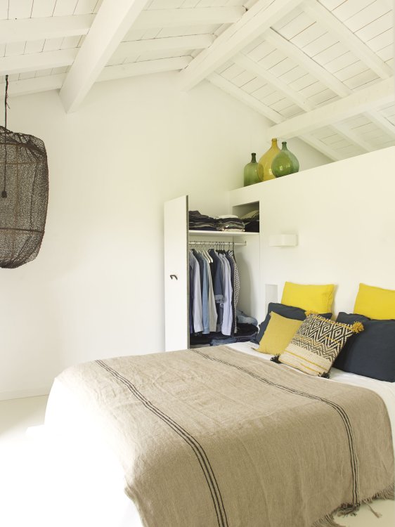 Ein Schlafzimmer im Boho-Chic-Style mit einem grossen Bett mit beiger Bettwäsche und Kissen in Gelb und Dunkelblau in einem weiss gestrichenen Dachbau.