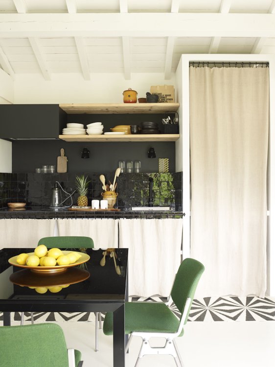 Ansicht einer Küche mit schwarzem Esstisch und grünen Stühlen im Vordergrund, dahinter eine kleine praktische Küche mit Vorratsschränken hinter hellen Leinenvorhängen.