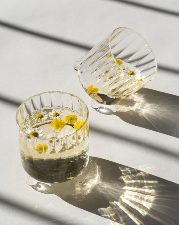 Zwei Trinkgläser, die je auf drei gläsernen Kugeln ruhen und ein Getränk mit weissen Blüten beinhalten, das hintere Glas ist leicht gekippt.