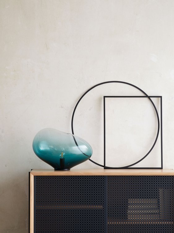 Eine organisch geformte Tischlampe aus Türkisen Glas steht auf einer schwarzen Kommode vor einem kreisrunden und rechteckigen Rahmenobjekt.