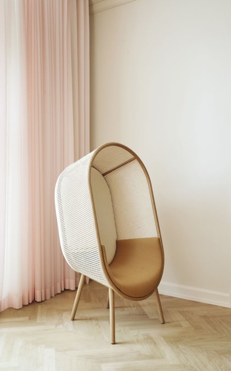 Ein moderner Loungesessel in Form eines Kokons aus Rattan und ockerfarbenem Polster steht in einer Ecke vor einem hellrosafarbenen Vorhang.