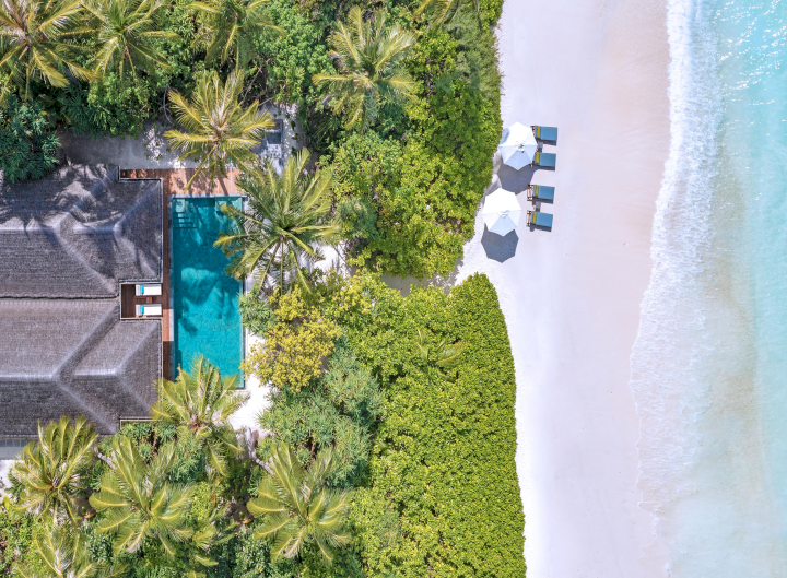 Eine Luftaufnahme einer exklusiven, von üppigem Grün umgebener Strandvilla des Anantara Kihavah Maldives Resorts mit einem kleinen geschlungenen Weg, der zu einem weissen Sandstrand mit Sonnenliegen direkt am türkisfarbenen Meer führt.