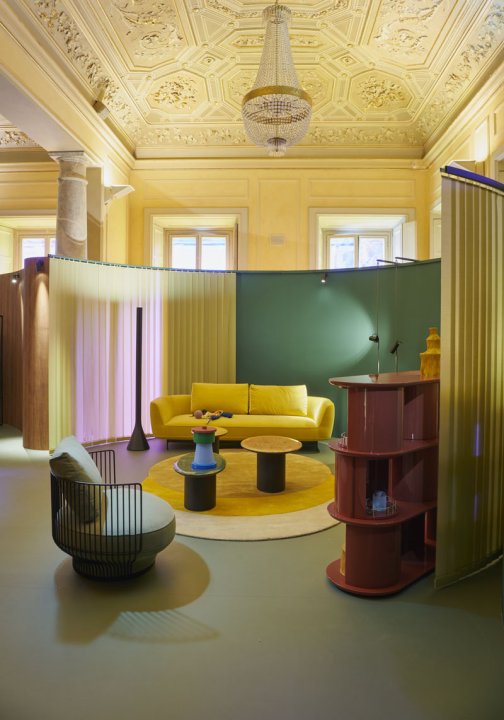 Die Installation von Wittmann während des Fuorisalone 2021 zeigt diverse bunte Möbel vor einer halbrunden Rückwand aus grünen, gelben, und pinken Stoffbahnen in einem gelben Salon eines Palazzos.