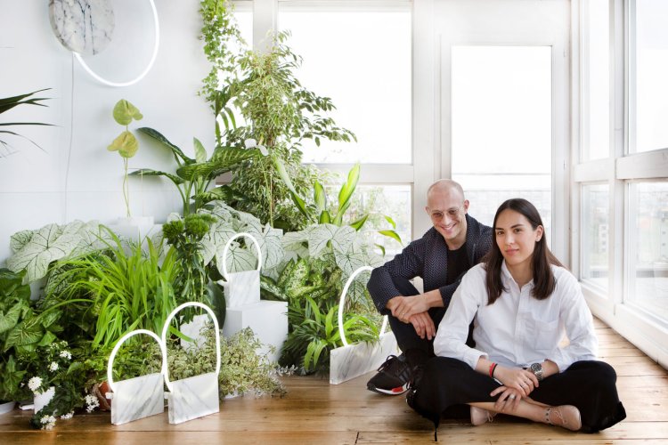 Porträt von Sara Ferron Cima, die gemeinsam mit Designer Carl Kleiner in einem Schneidersitz am Boden sitzt, links im Raum steht eine grosse Menge grüner Pflanzen, davor sind einige Lampen mit Marmorsockel von ihrem Studio Bloc aufgestellt.
