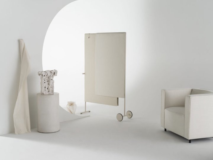 Ein weisses Setting in dem eine beige Trennwand auf Rollen neben einem kubisch geformten Sessel neben weiteren weissen und hellen Objekten stehen.