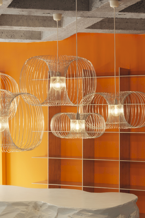Diverse Varianten der Coil-Hängelamoe aus abgerundeten Edelstahldrähten hängen in einem Raum mit knallorangen Wänden.
