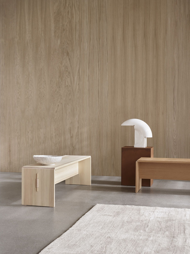 Zei Bänke in schlichtem Design und in hellem und dunklerem Holz stehen in einem minimalistisch eingerichteten Raum mit einem hellen Teppich und einer Wand mit Holzverkleidung.