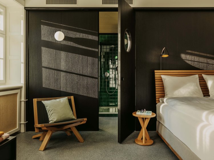 Ein Gästezimmer im Boutique Hotel Volkshaus mit einem Doppelbett und Loungechair vor einer schwarzen Holzwand, hinter der das Badezimmer mit dunkelgrünen Kacheln sichtbar ist.