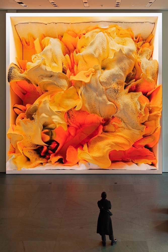 Eine Besucherin steht vor einer quadratischen Bewegtbild-Installation in orange und gelben Farbwolken im Entree des Museum of Modern Art in New York.