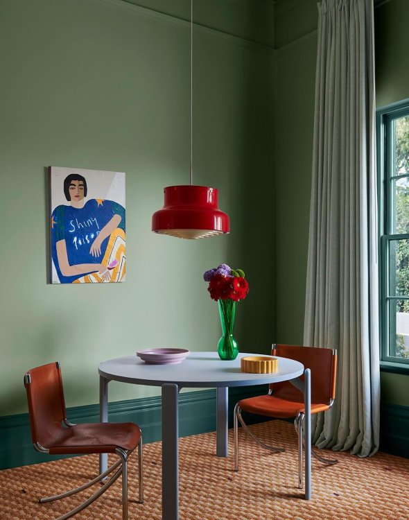 Ein olivgrün gestrichenes Wohnzimmer mit einem hellblauen runden Tisch an dem zwei Freischwinger Stühle in rostrot stehen, darüber hängt eine Pendelleuchte in knalligem Rot.
