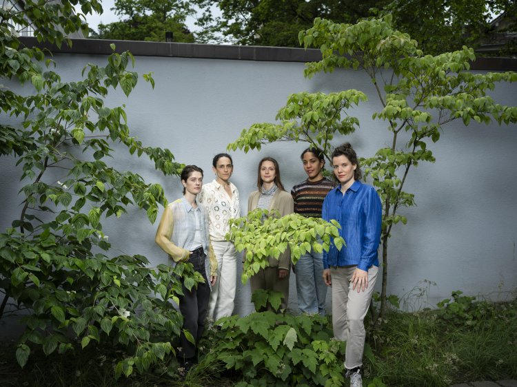 Ein Porträt von Axelle Stiefel, Elena Chiavi, Amy Perkins, Myriam Uzor und Kathrin Füglister vor einer blauen Wand, umgeben von grünen Pflanzen.