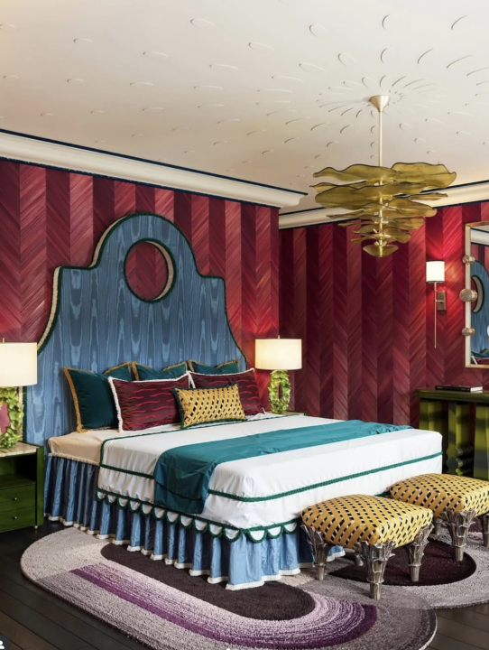 Ein Schlafzimmer mit kunterbunter Einrichtung, rot gestreifter Tapete, blauem Kopfteil und bunt gemischten Kissen auf einem organisch geformten Teppich in Violett-Tönen.