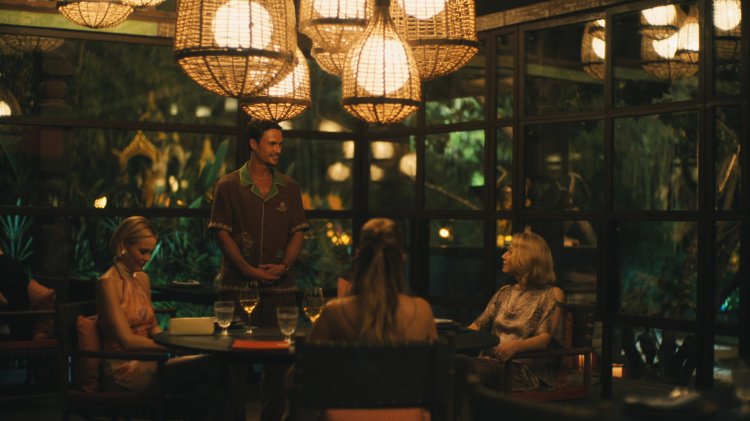 Eine Szene aus The White Lotus Staffel 3 zeigt drei Freundinnen beim Outdoor Dinner unter geflochtenen Lampen.