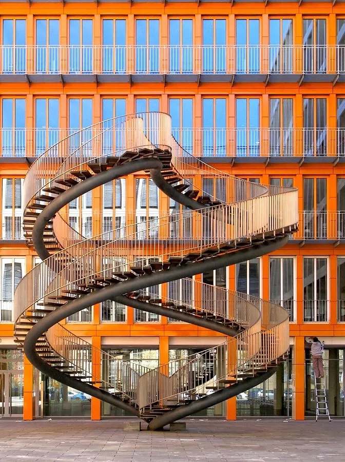Freistehende Aussenskulptur aus silbergrauem Stahl in Form einer Doppelspirale vor einem orangen Bürogebäude.