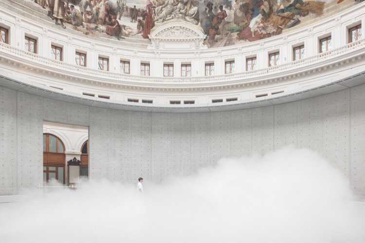 In der runden Halle der Bourse de Commerce liegt dichter weisser Nebel am Boden und eine einzelne Person steht bis zu Schulterhöhe darin verschwunden.