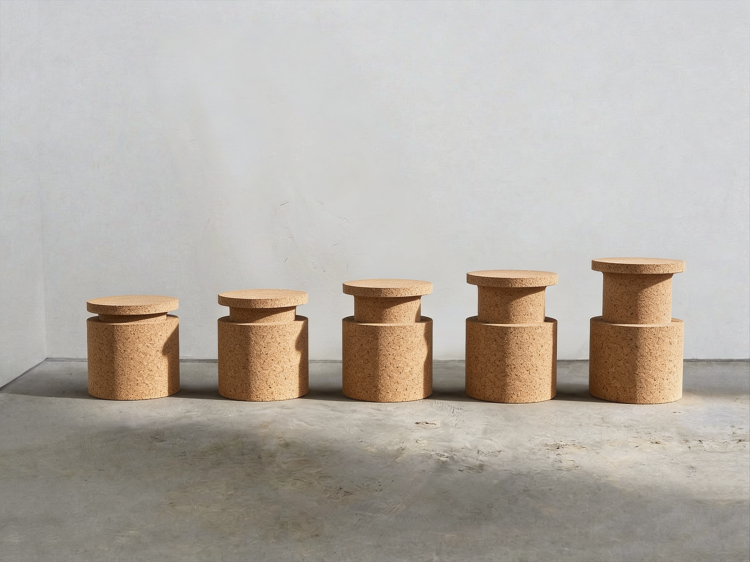 Fünf zylindrische Hocker aus hellem, körnigem Kork stehen nebeneinander vor einer weissen Wand, aufsteigend von niedrig bis hoch.