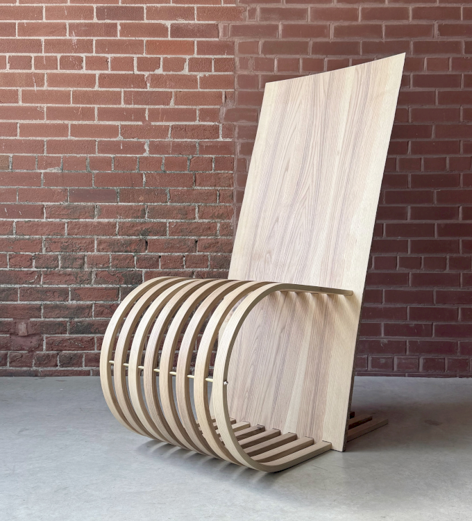 Ein Loungesessel aus hellem Holz mit geschwungener Massivholzlehne und gerippter Sitzfläche steht vor einer Backsteinwand.