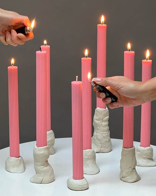 Mehrere rosa Kerzen, die bereits brennen oder von zwei Händen bestückt mit Feuerzeug angezündet werden.
